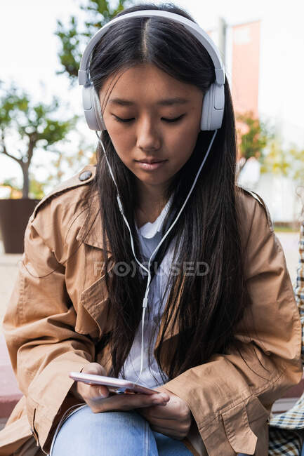 Giovane focalizzata femminile asiatica in abiti casual seduto sulla strada con cuffie navigazione internet sul telefono cellulare — Foto stock