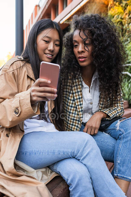 Pleasant Asian female with toothy smile showing video on smartphone to happy black woman while spending time together — Stock Photo