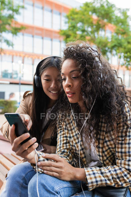 Asian female having fun call on smartphone and focused black woman listening to music and surfing internet on mobile phone — Fotografia de Stock