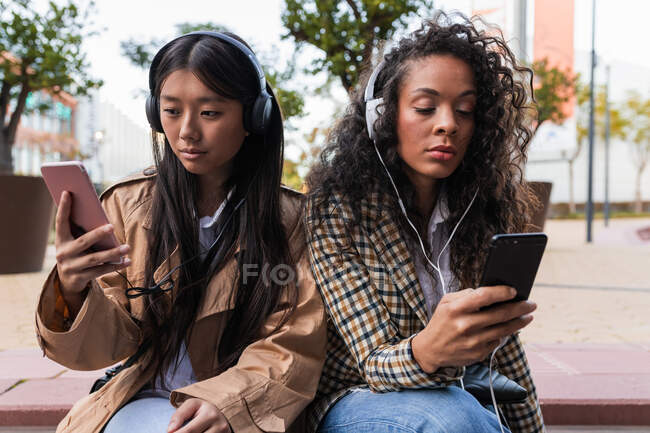 Pensativo asiático hembra divertirse llamada en el teléfono inteligente y centrado negro mujer escuchando música y navegar por Internet en el teléfono móvil - foto de stock