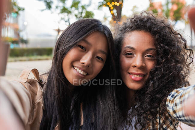 Delighted Asian female and positive black woman in casual clothes looking at camera while taking selfie on smartphone — Photo de stock