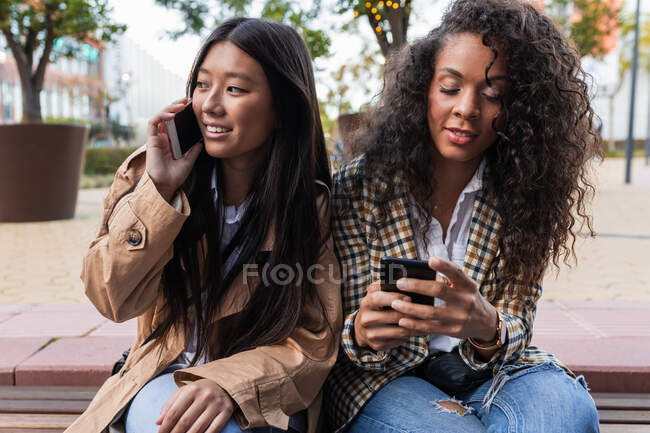 Happy asian female having fun call on smartphone and black woman surfing internet on mobile phone — Fotografia de Stock