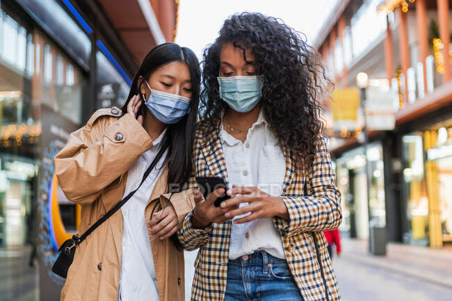 Mujeres étnicas jóvenes con máscaras protectoras que usan ropa casual de pie en la calle de la ciudad y navegando por el teléfono móvil mientras encuentran la dirección - foto de stock