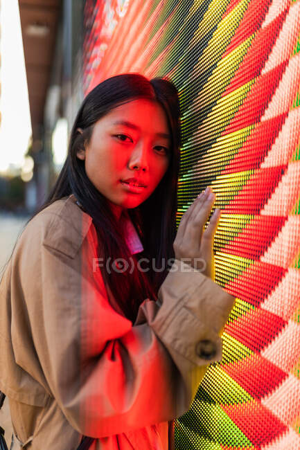 Wistful ethnic female in casual clothes with long hair looking at camera while leaning on wall with neon illumination — Stock Photo