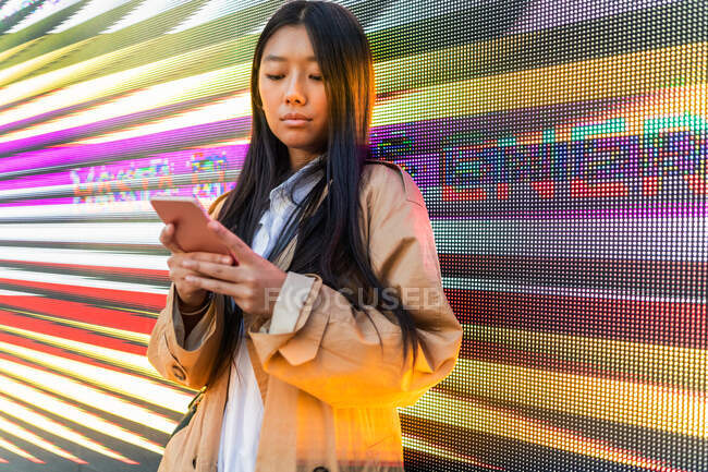 Серйозна азіатка в модному вбранні перегляд мобільного телефону проти цифрової стіни — стокове фото