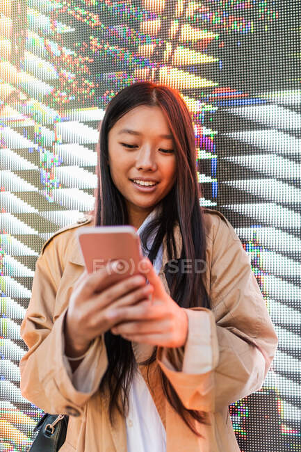 Happy asian female in trendy outfit browsing mobile phone against digital wall — Photo de stock