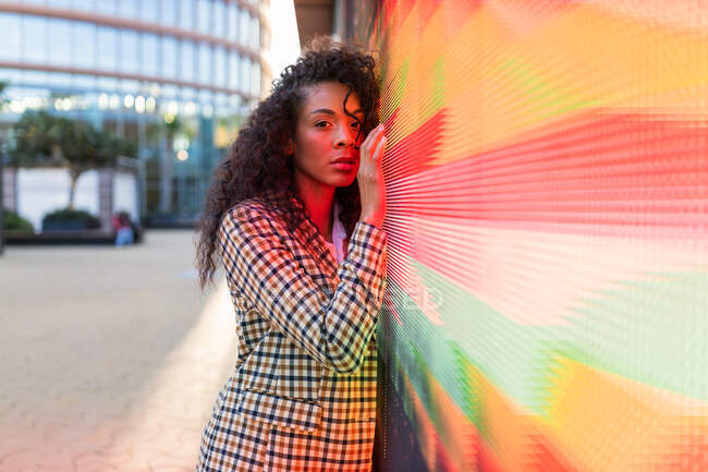 Scharfsinnige schwarze Frau in lässiger Kleidung mit lockigem Haar blickt in die Kamera, während sie sich an eine Wand mit Neonbeleuchtung lehnt — Stockfoto
