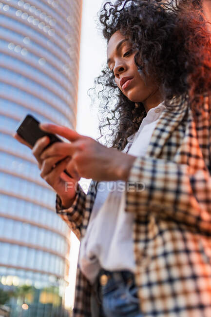 Low angle of thoughtful African American female with curly hair standing on street and using mobile phone for checking social networks — Photo de stock
