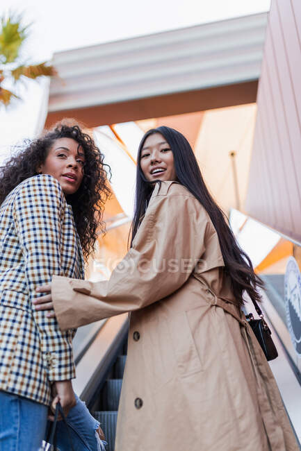 Óptima hembra asiática mirando a la cámara mientras camina con elegante mujer negra en el centro de la ciudad - foto de stock