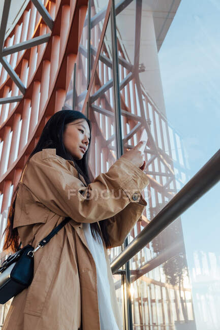 From below of serious asian woman in stylish outfit looking out window while taking picture on mobile phone — Photo de stock