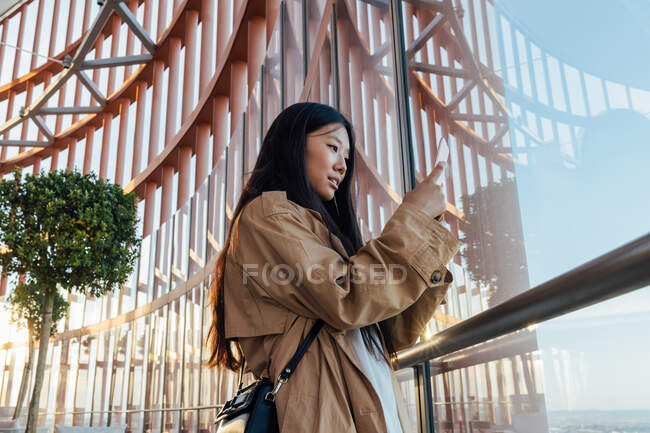 Side view of serious asian woman in stylish outfit looking out window while taking picture on mobile phone — Photo de stock