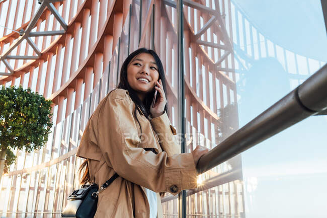 Seitenansicht einer glücklichen Asiatin im stylischen Outfit, die wegschaut, während sie auf dem Handy spricht — Stockfoto