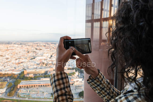 Vista trasera de la mujer negra en traje elegante mirando por la ventana mientras toma la foto en el teléfono móvil - foto de stock