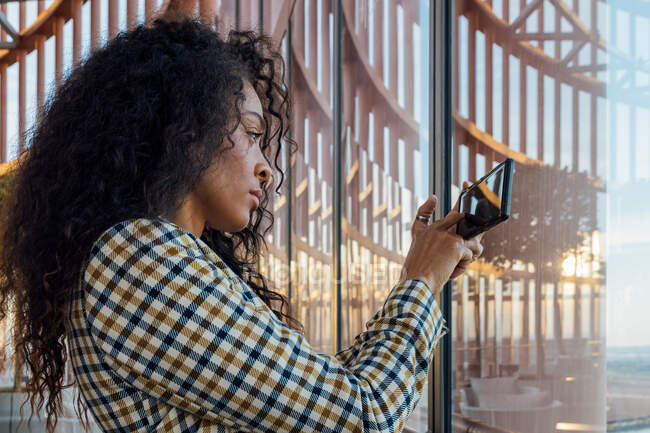 Vista lateral de una mujer negra seria con un atuendo elegante mirando por la ventana mientras toma fotos en el teléfono móvil - foto de stock