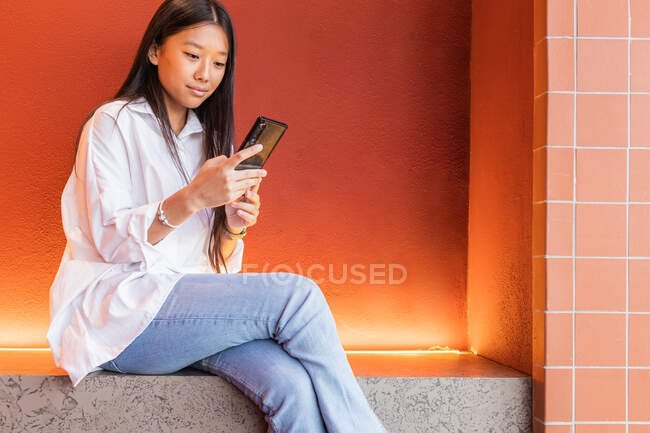 Спокійна азійка у повсякденному одязі сидить у зручній позі і переглядає Інтернет на мобільному телефоні. — стокове фото