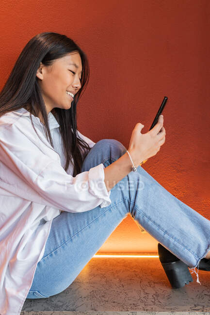 Happy asian woman in casual clothes sitting in comfortable pose and browsing internet on mobile phone — Fotografia de Stock