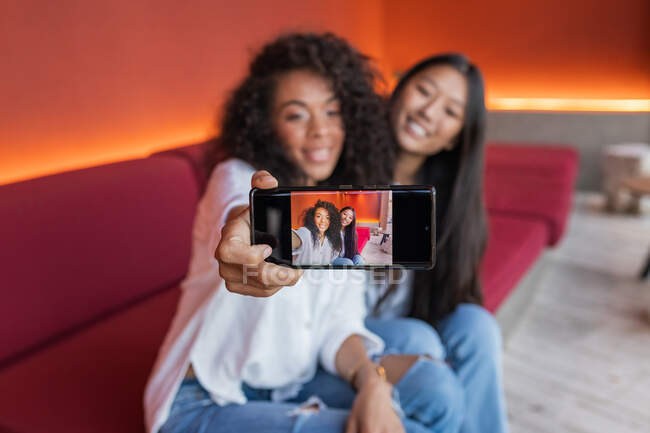 Young cheerful black female and satisfied Asian girlfriend sitting on sofa and taking selfie on mobile phone while resting — Photo de stock