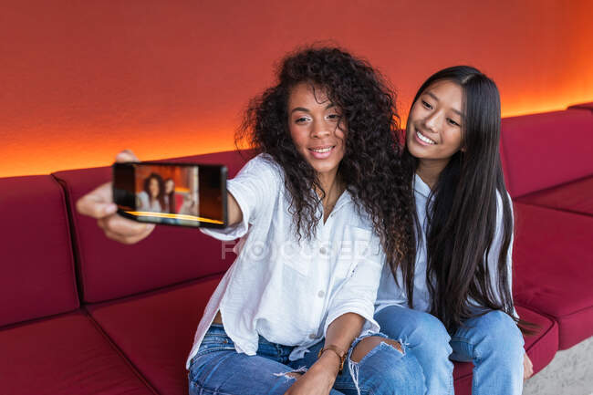 Молодая веселая черная женщина и довольная азиатская девушка сидит на диване и делает селфи на мобильном телефоне во время отдыха — стоковое фото
