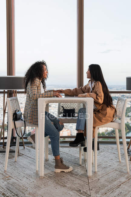 Вид сбоку на стильных восторженных многоэтнических подруг, говорящих и сидящих за соседним столиком в кафе — стоковое фото
