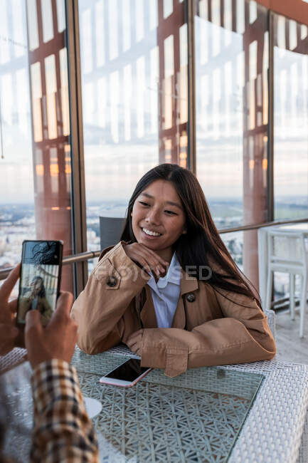 Crop person taking photo of positive Asian woman with toothy smile sitting at table in light cafeteria — Fotografia de Stock