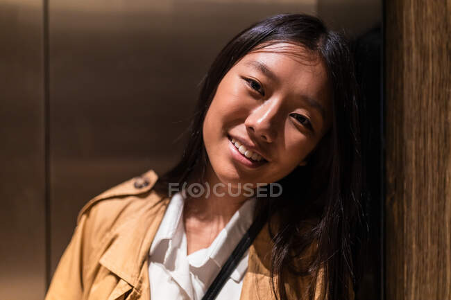 Retrato de mujer asiática optimista en ropa casual con sonrisa dentada mirando a la cámara mientras se apoya en la pared de madera - foto de stock