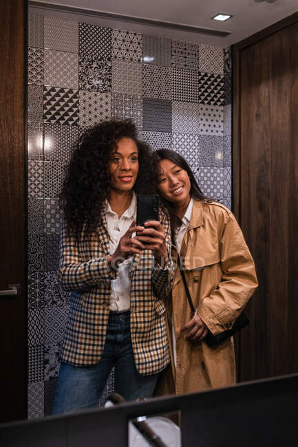 Young African American woman in stylish clothes and positive Asian female looking in mirror while taking selfie on mobile phone — Photo de stock