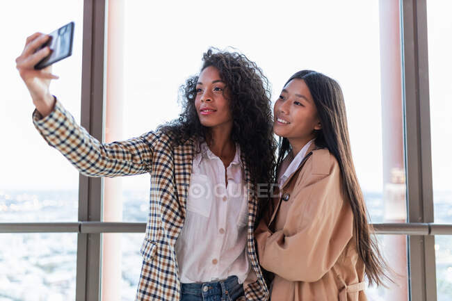 Young cheerful black female and satisfied Asian girlfriend standing in cafe and taking selfie on mobile phone — Photo de stock
