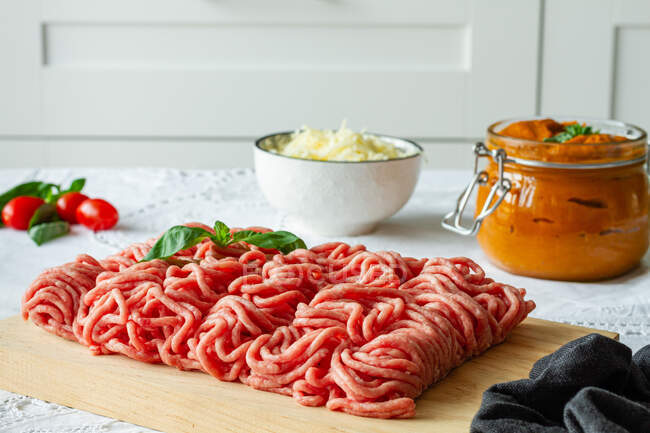 From above of fresh raw mixed pork and beef minced meat on wooden cutting board with green basil leaves placed on table during cooking process near homemade tomato sauce — Stock Photo