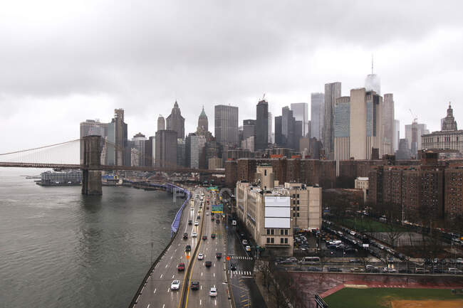 Район Манхэттена с городскими небоскребами и подвесным мостом над бурлящей Ист-Ривер под мрачным облачным небом — стоковое фото