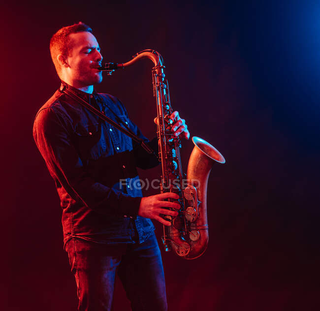 Профессиональный музыкант с закрытыми глазами играет на саксофоне в красном и синем неоновом свете во время живого выступления — стоковое фото