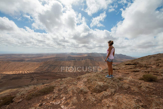 Back view anonymous female hiker in casual outfit standing on rocky hill and enjoying spacious hilly valley on clear day — Fotografia de Stock