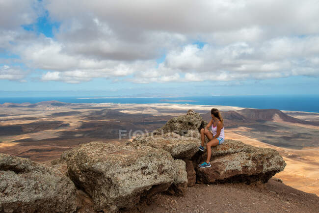 Боковой вид женщины-туристки в повседневной одежде сидит на скалистом холме и наслаждается просторной холмистой долиной в ясный день — стоковое фото
