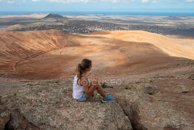 Caminante femenina vista lateral en traje casual sentada en una colina rocosa y disfrutando de un amplio valle montañoso en un día despejado - foto de stock