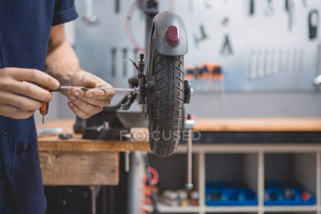 Side view of crop anonymous male mechanic with screwdriver fixing wheel of electric scooter in workshop — Fotografia de Stock