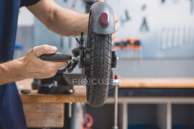 Side view of crop anonymous male mechanic with screwdriver fixing wheel of electric scooter in workshop — Stock Photo