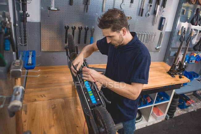 Вид сбоку на концентрированного квалифицированного механика-мужчину с помощью мультиметра при тестировании аккумулятора электрического скутера в мастерской — стоковое фото