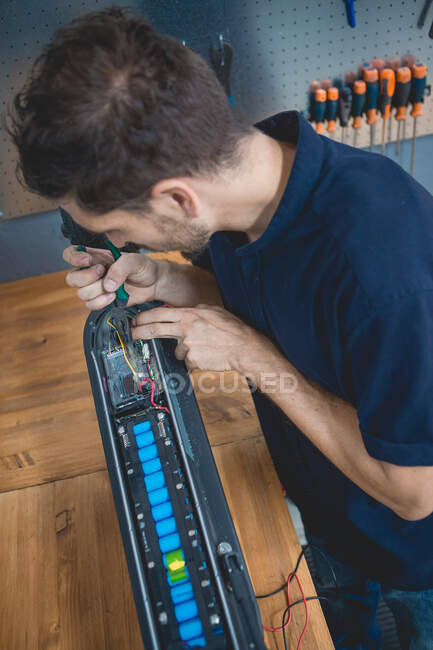 Vista lateral del mecánico masculino experto concentrado usando multímetro mientras prueba la batería de scooter eléctrico en el taller - foto de stock