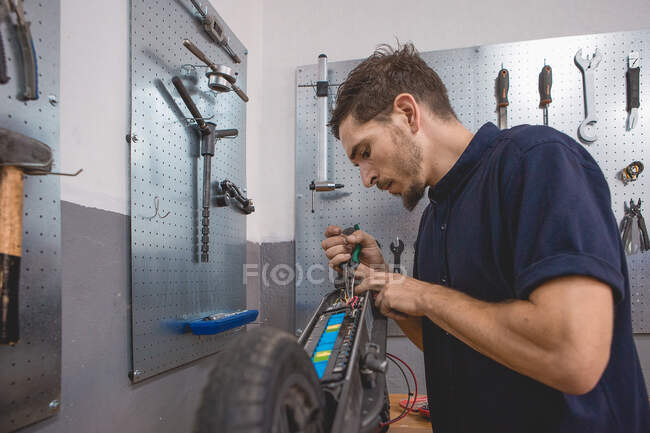 Vista lateral del mecánico masculino experto concentrado usando multímetro mientras prueba la batería de scooter eléctrico en el taller - foto de stock