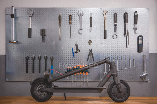 Scooter eléctrico incompleto colocado cerca de la pared con varias herramientas e instrumentos de reparación en el taller moderno - foto de stock