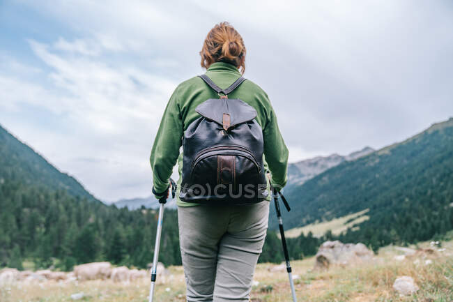 Back view of anonymous female hiker in casual clothes with backpack and Nordic walk poles standing in picturesque highlands — Stock Photo