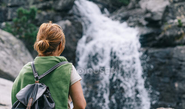 Вид на анонимную женщину-туристку в повседневной одежде с рюкзаком, стоящим в лесу и любующимся видом на мощный водопад — стоковое фото