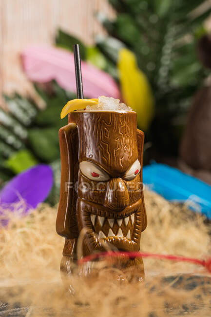 Brown sculptural tiki mug with alcohol drink decorated with straw and ice placed amidst dry grass against green plant with feather on blurred background — Stock Photo