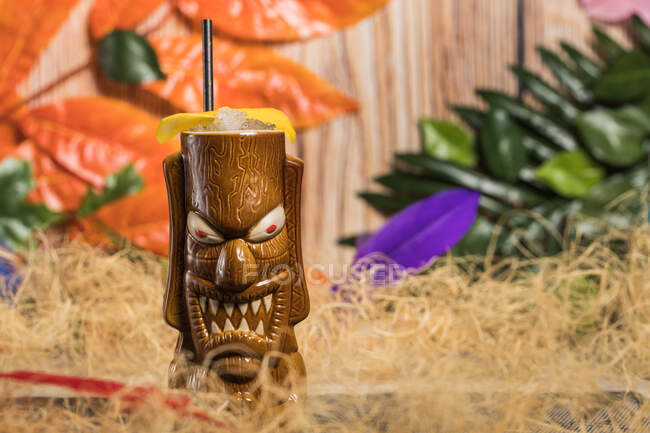 Tasse à tiki sculpturale marron avec boisson alcoolisée décorée de paille et de glace placée au milieu de l'herbe sèche contre une plante verte avec plume sur fond flou — Photo de stock