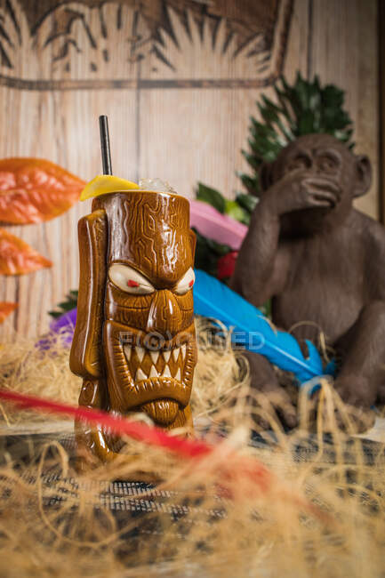 Brauner skulpturaler Tiki-Becher mit Alkoholgetränk verziert mit Stroh und Eis inmitten von trockenem Gras vor grünem Gewächs mit Feder auf verschwommenem Hintergrund — Stockfoto