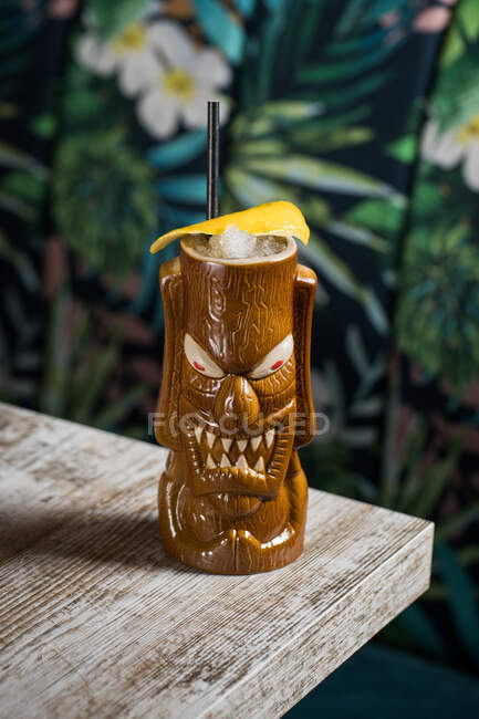 Tazza di tiki scultorea marrone con bevanda alcolica decorata con paglia e ghiaccio posta sul tavolo su sfondo sfocato — Foto stock