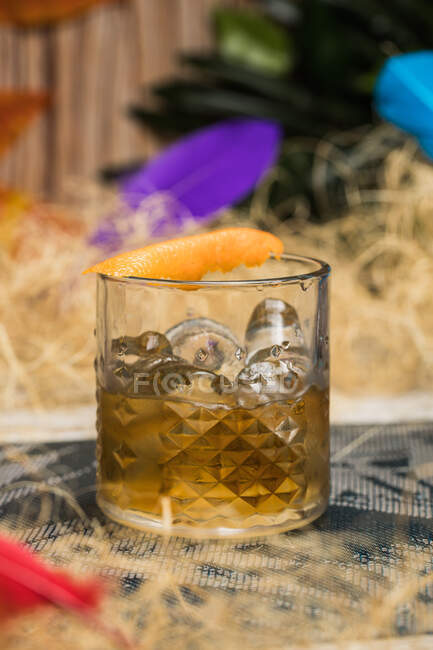 Tasse en verre Tiki avec boisson à l'ancienne placée sur un tissu au milieu de l'herbe sèche contre une clôture en bois et des feuilles colorées sur fond flou — Photo de stock