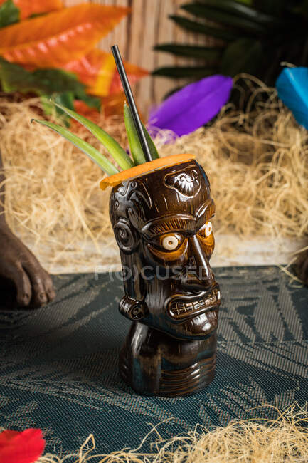 Ceramic polynesian mug with alcohol drink served with straw and decorations placed on cloth against dry grass and colorful leaves — Stock Photo