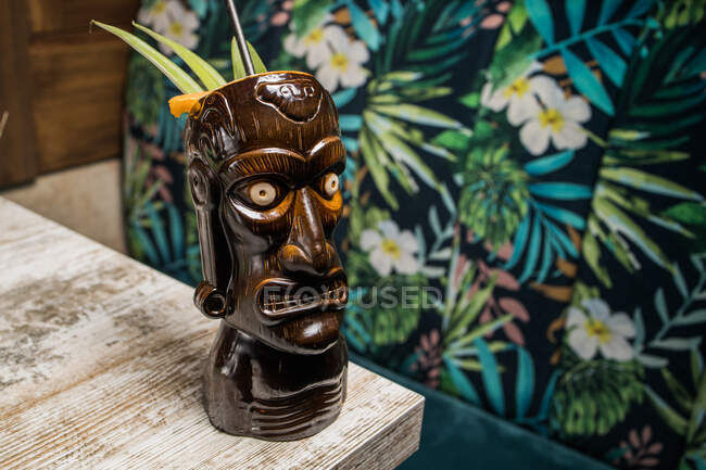 Taza tiki escultórica marrón con bebida alcohólica decorada con paja y hielo colocada en una mesa de madera - foto de stock