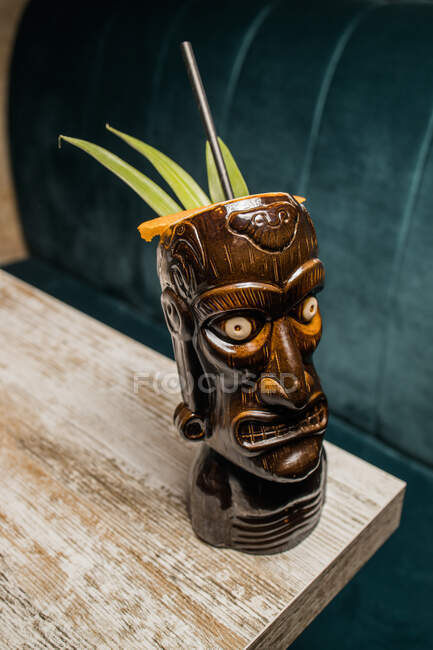 Tasse à tiki sculpturale marron avec boisson alcoolisée décorée de paille et de glace placée sur une table en bois — Photo de stock