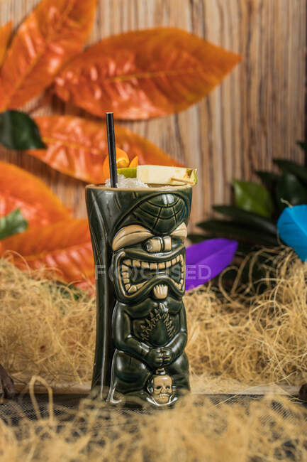 Large sculptural tiki cup filled with booze decorated with straw and fruits placed on green rug against dry grass — Stock Photo
